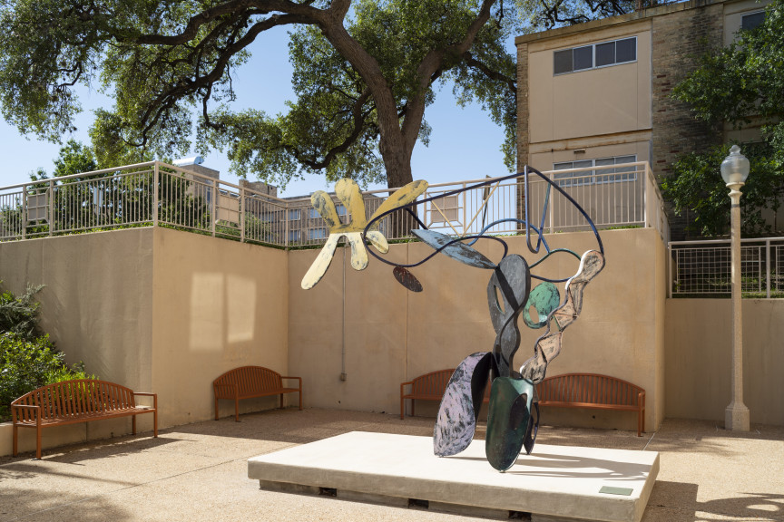 A large bendy sculpture with parts colored in different colors in a courtyard with benches surrounding it