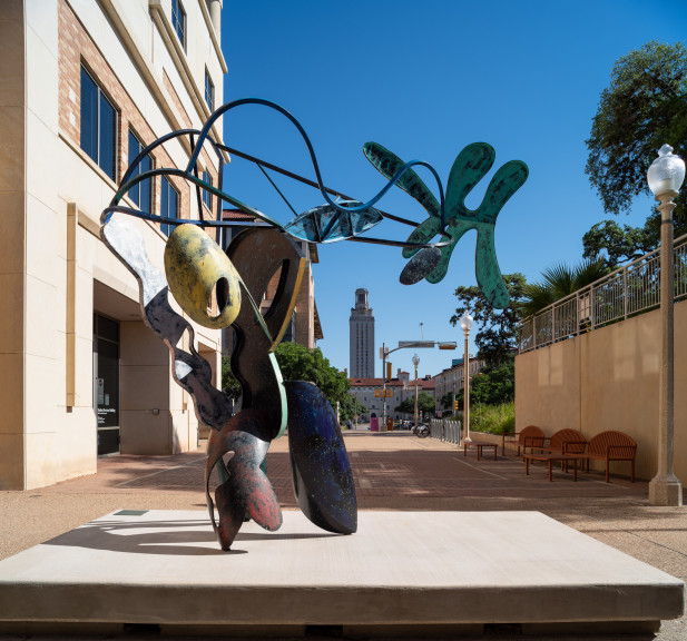 A large bendy sculpture with parts colored in different colors in a courtyard as seen from behind with the UT Tower framed by the sculpture