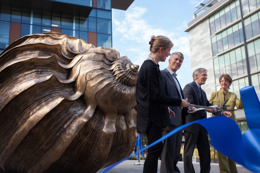 Four people cutting a large ribbon in front of a bronze shell