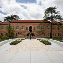 A wide angle image of Simone Leigh's Sentinel IV in the courtyard of Anna Hiss Gym. The building seems to surround the image and has a central staircase in the center of the image. The sculpture, a slender bronze figure with a bowllike head is placed in the center.
