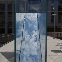 An image of Sarah Oppenheimer's "C-010106," two works cut through the surface of a pedestrian bridge. In this image, a blue sky with clouds is reflected on the diagonal surface of the work.