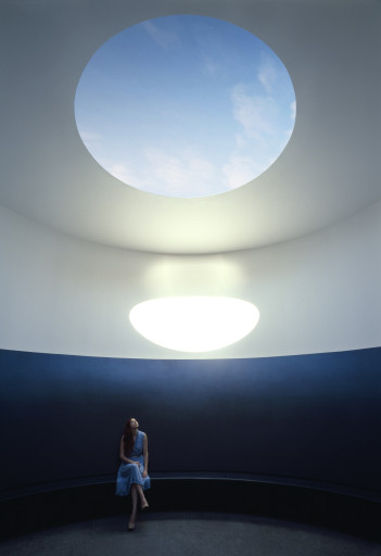 A woman sits in James Turrell's Skyspace, "The Color Inside" during the day. The sun shines through a hole in the ceiling.