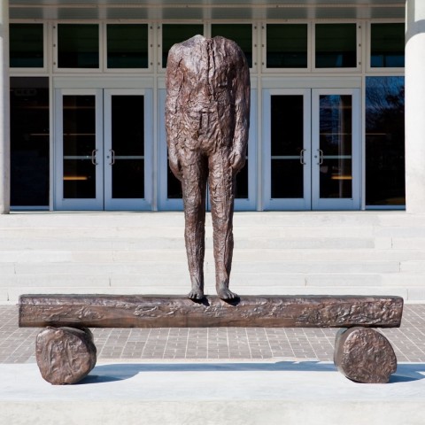 A headless figure stands on a flat surface supported by two wheels which look like tree trunks. The work is cast bronze and hollow when viewed from the other side
