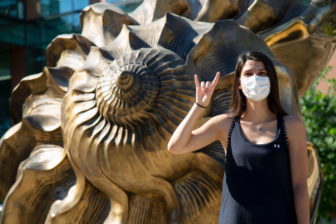 A woman in a mask makes a "hook em" gesture and looks at the camera in front of Marc Quinn's sculpture "Spiral of the Galaxy" which is an enlarged bronze seashell