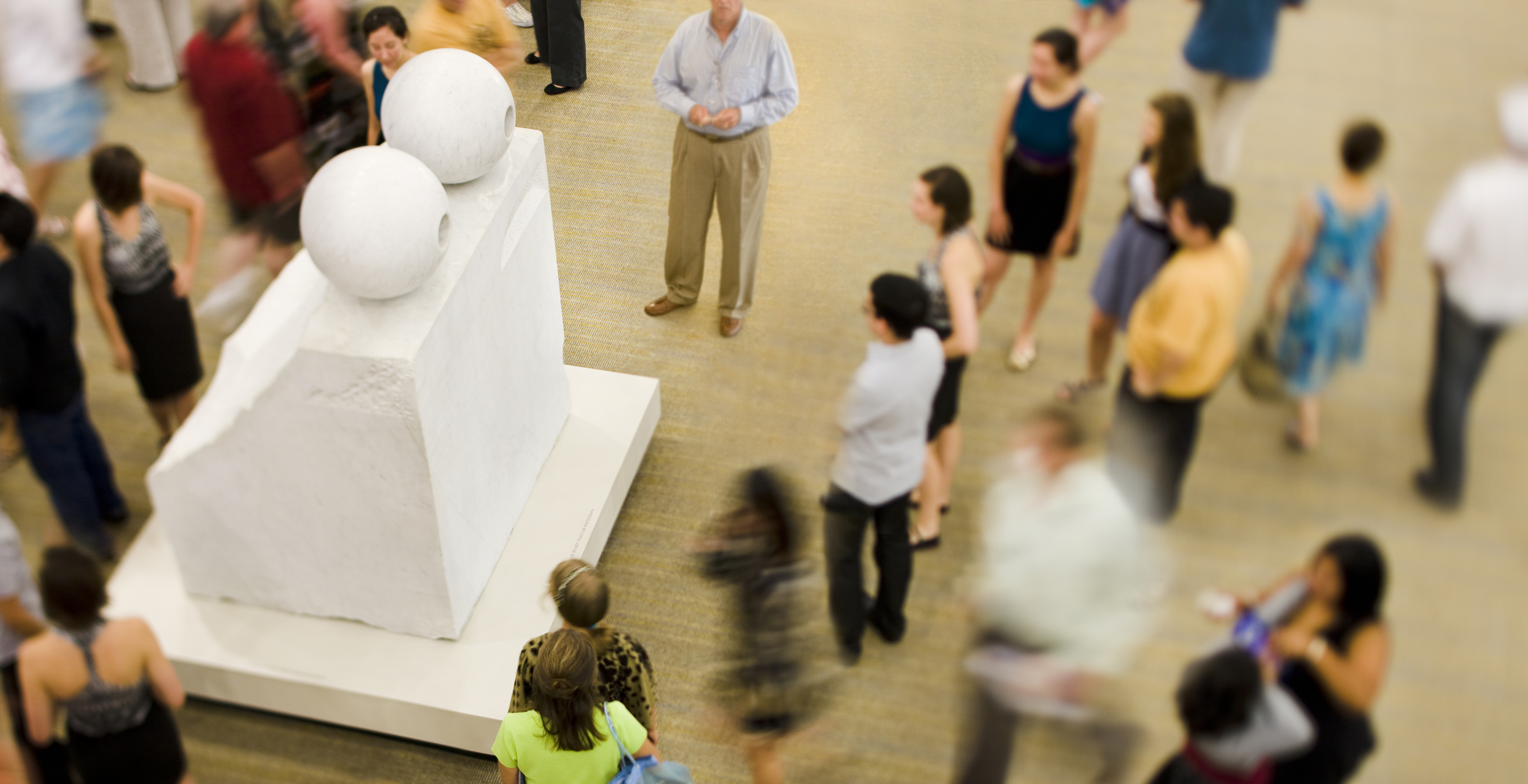 A group of people seen from above surrounding Louise Bourgeois' Sculpture "Eyes" a white marble sculpture that is a block with two circular eyes carved on top. 