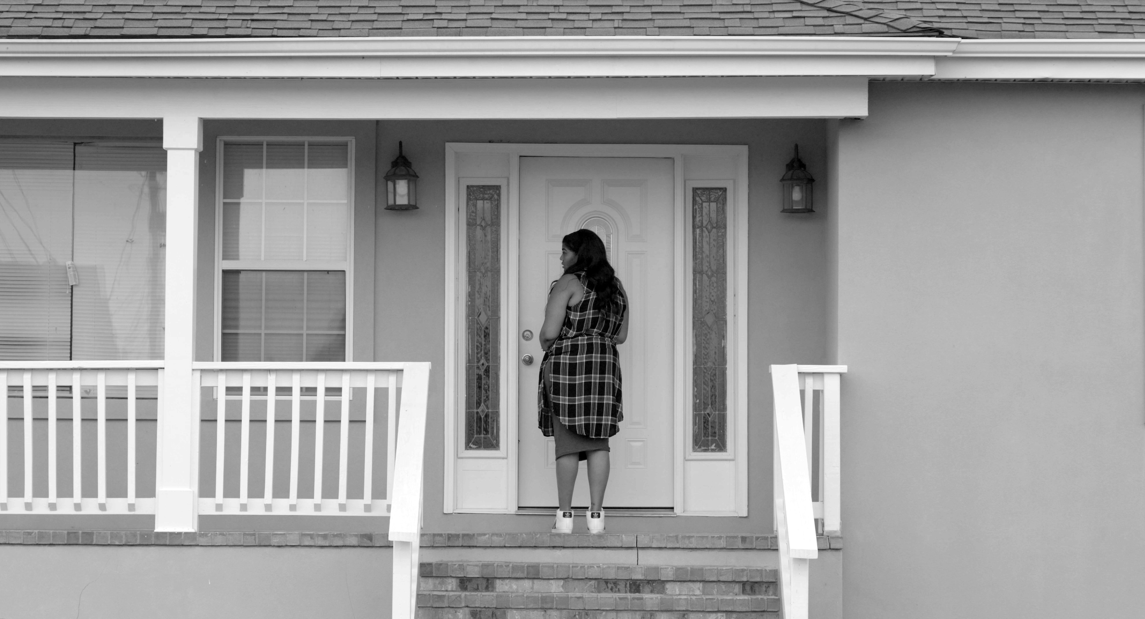 A still from Garrett Bradley's video "Alone" which shows a front porch with a female figure standing at a front door. The entire scene is in grey-scale. 