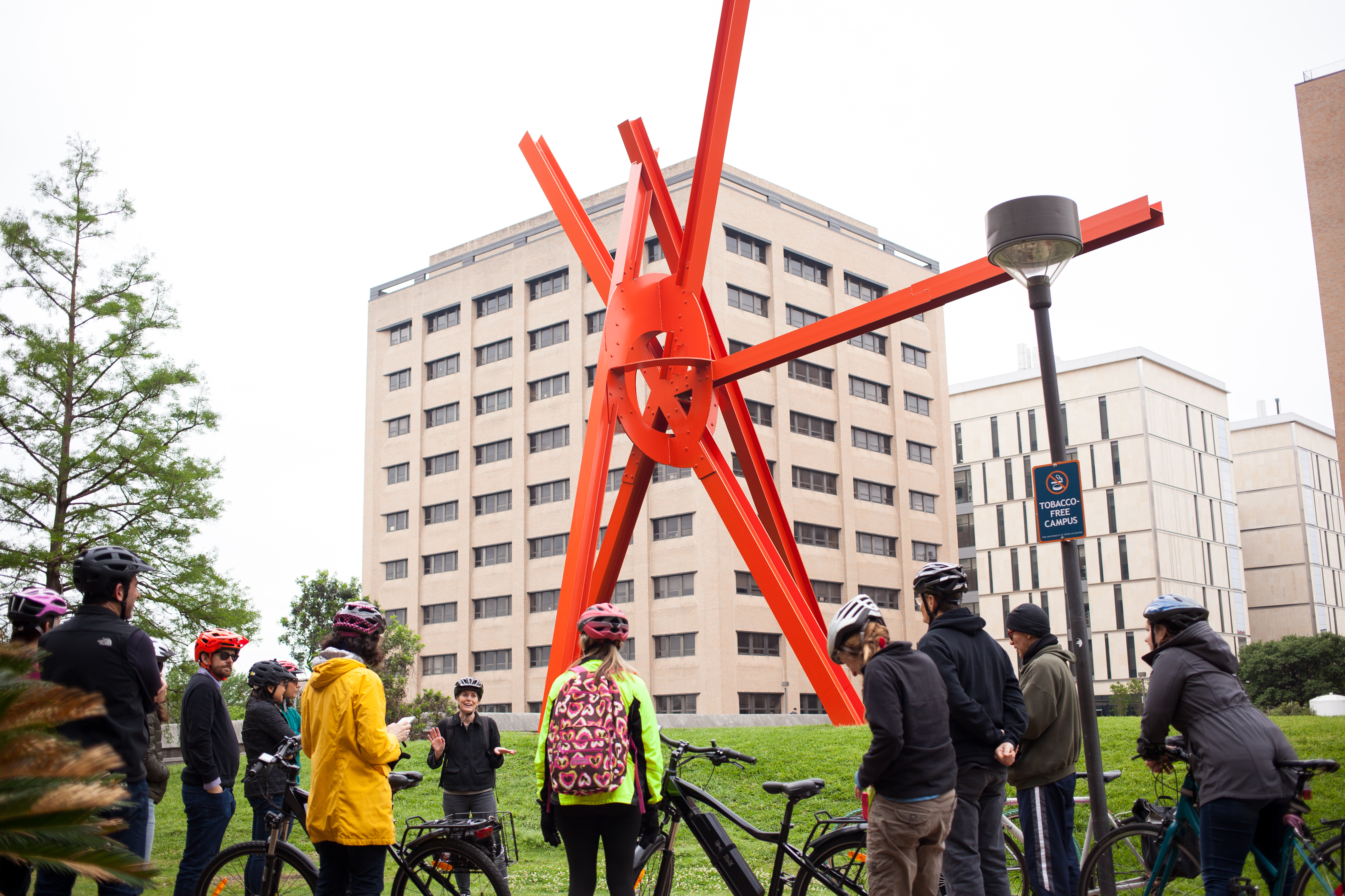 A group of people with bikes wear helmets and stand looking at Marc di Suvero's "Clock Knot," a large red sculpture that juts out over the green hill it's placed on 