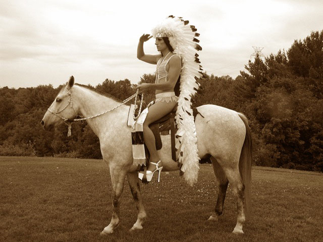 A man in a long Native American headdress and large pumps sitting on a horse