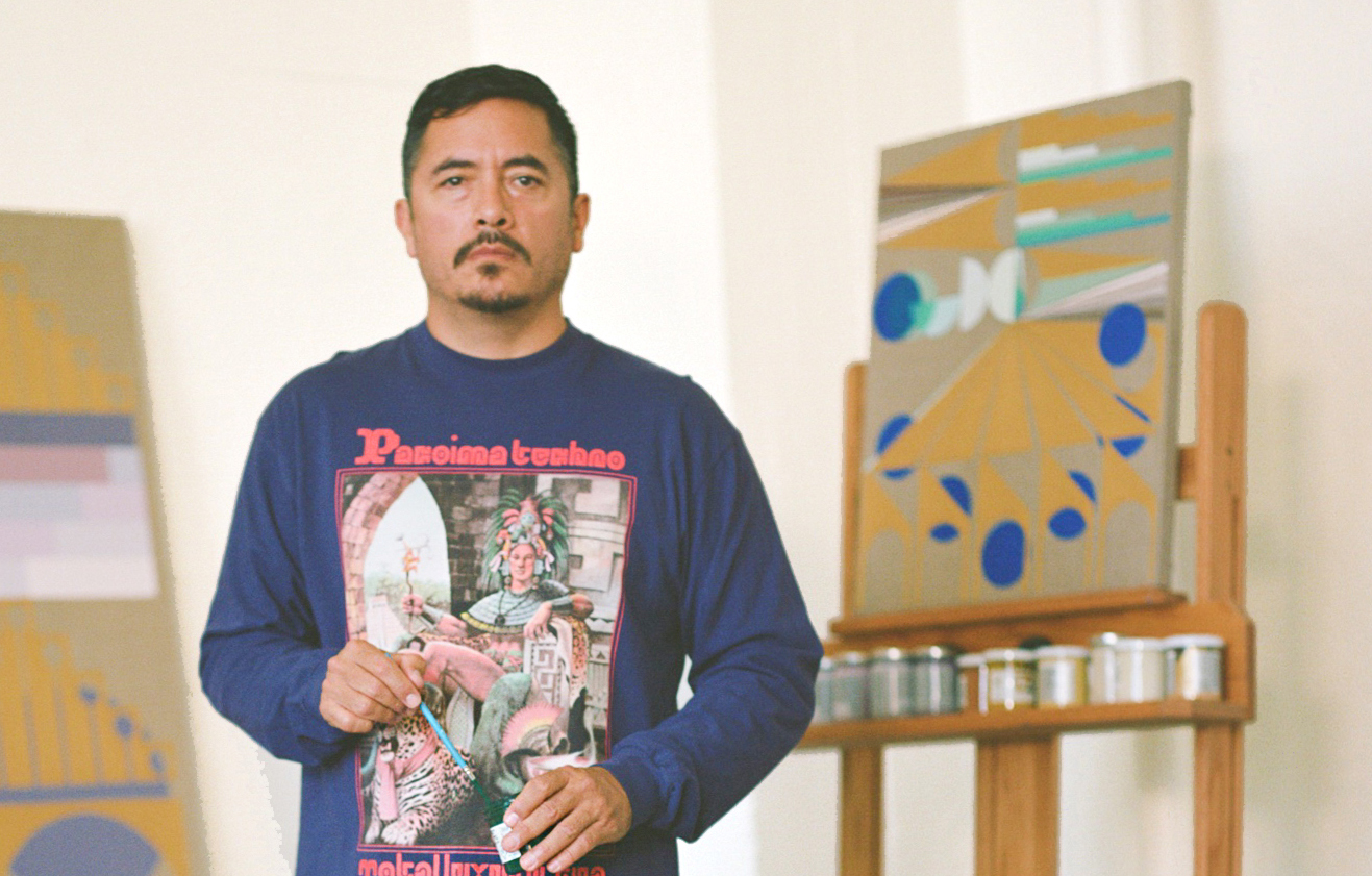 An image of Eamon Ore-Giron who stands with a paintbrush in hand and in front of his paintings