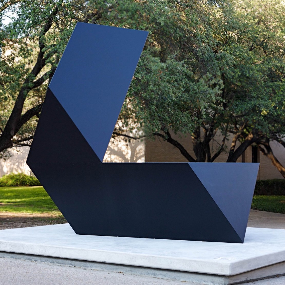 A deep black sculpture composed of elongated, triangular sections. In this view the sculpture forms an "L" shape