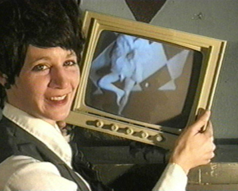 A short haired woman in a vest holding a black and white television