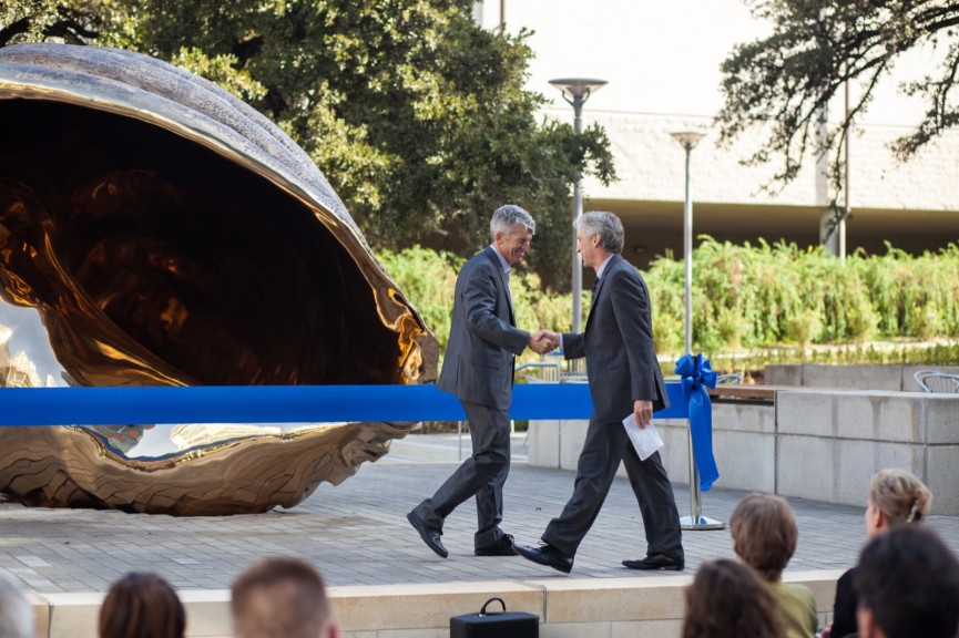 Two people shaking hands in front of a large ribbon and bronze shell