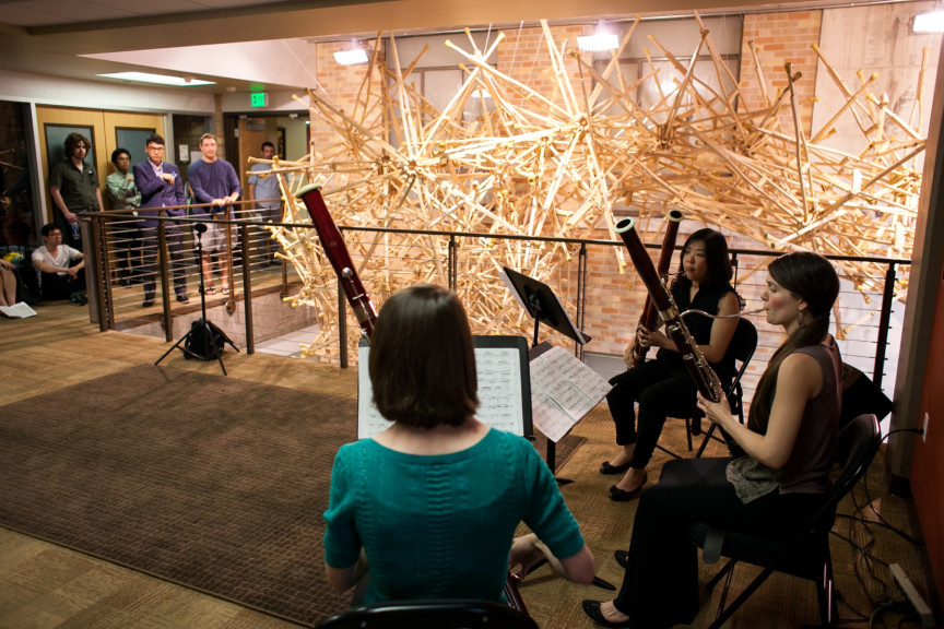 People performing with oboes next to a hanging sculpture made out of wooden crutches