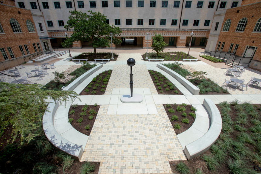 A wide angle image of Simone Leigh's Sentinel IV in the courtyard of Anna Hiss Gym. The building seems to surround the image and has a central staircase in the center of the image. The sculpture, a slender bronze figure with a bowllike head is placed in the center.