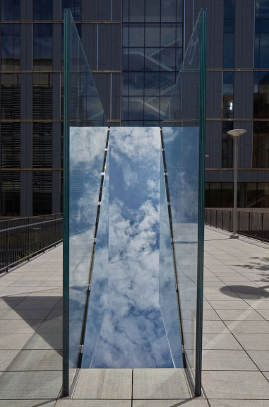 An image of Sarah Oppenheimer's "C-010106" which shows a blue sky with clouds reflected in the work.