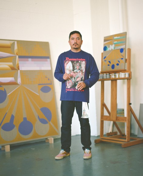 An image of Eamon Ore-Giron standing in his studio. He holds a paintbrush and stands in front of in-progress paintings