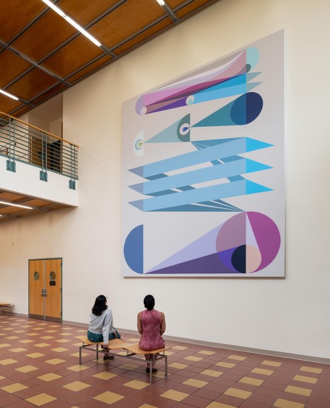 An image of Eamon Ore-Giron's "Tras los ojos," a large digital print which hangs in the atrium of a building. This images includes two women who sit on a bench in front of the work.