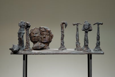 Gray sculptural assemblage on a narrow table, featuring several upright, tool-like forms and a cluster of three faces at the center.