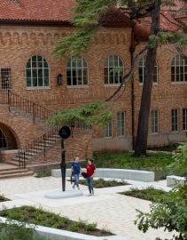 Two people seen from a high vantage point look at Simone Leigh's Sentinel IV, a tall slender figure with a bowlcrown in place of a head. They are surrounded by architecture and the landscaping of the space. 