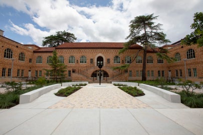 A wide angle image of Simone Leigh's Sentinel IV in the courtyard of Anna Hiss Gym. The building seems to surround the image and has a central staircase in the center of the image. The sculpture, a slender bronze figure with a bowllike head is placed in the center.