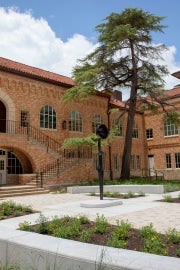 The artwork, Sentinel IV, a tall slender female figure with a bowlcrown for a head is centrally placed in the courtyard of Anna Hiss Gymnasium. There is a large tree that can be seen to the right of the image. 