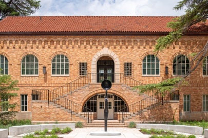 Simone Leigh's Sentinel IV, a tall slender female figure is seen from straight on in the courtyard of Anna Hiss Gymnasium. The work is framed by a large staircase which goes up left and right behind the sculpture. The red/orange brick of the building stands out against the bright blue sky.  