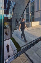 A detail of Sarah Oppenheimer's "C-010106," which shows the reflective quality of the work. Three people are caught in motion through the reflection of the work.
