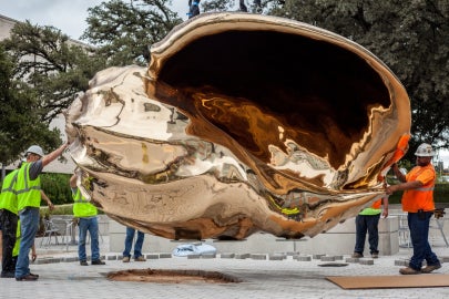 A bronze shell hanging from a crane inches above the ground with crew members surrounding it