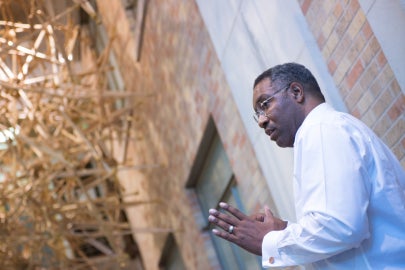 A man in a collared shirt standing underneath a hanging sculpture made of crutches