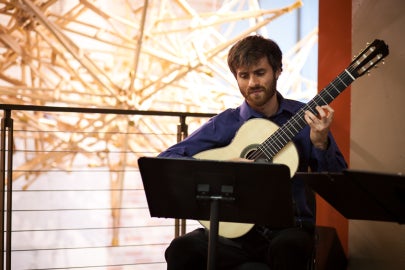 A man playing an acoustic guitar next to a hanging sculpture made out of wooden crutches