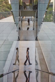 A detail of Sarah Oppenheimer's "C-010106," which shows the reflective quality of the work. Three people are caught in motion through the reflection of the work.