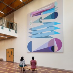 An image of Eamon Ore-Giron's "Tras los ojos," a large digital print which hangs in the atrium of a building. Two women sit on a bench in front of the work.