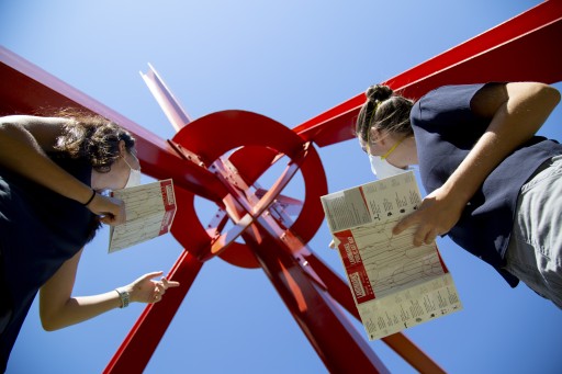 Two women in masks holding Landmarks maps look up at Mark Di Suvero's sculpture "Clock Knot"