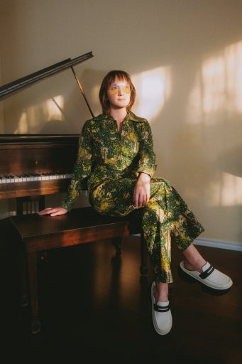 a woman with short brown hair, yellow aviators, and a green patterned jumpsuit sits at a piano.