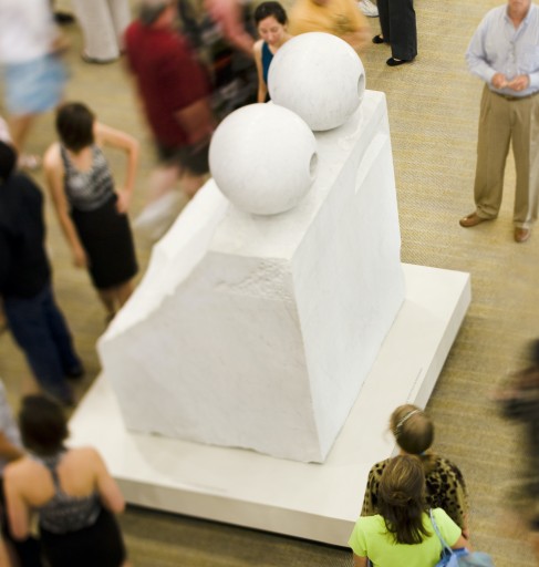 An image of Louise Bourgeois' sculpture "Eyes" a block of white marble with two circular carved eyes perched on top. The work is seen from above and is surrounded by a crowd of people who look and move around it