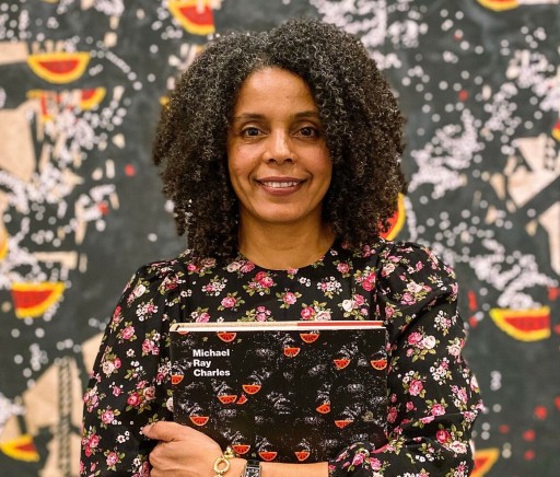 An image of Dr. Cherise Smith holding a copy of her book "Michael Ray Charles: A Retrospective" the pattern of the book is repeated in the background of the image
