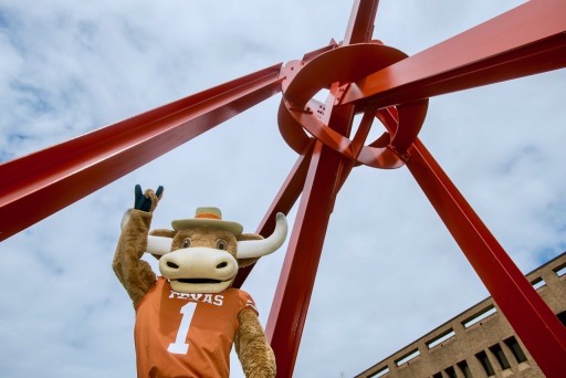 "Hook Em" UT's Longhorn Mascot, holds up a "hook em" gesture in front of Mark di Suvero's "Clock Knot" a large red metal sculpture that seems to meet in a mass in the upper right corner of the image