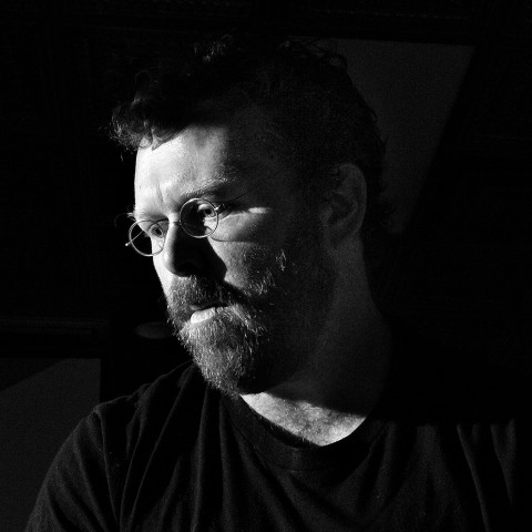 A black and white headshot of a man with a beard and glasses. 