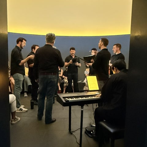 Arranged in a tight circle, a group of choral singers preform with a keyboard player.