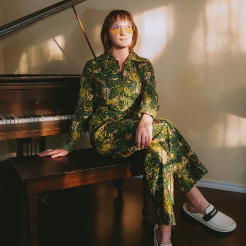a woman with short brown hair, yellow aviators, and a green patterned jumpsuit sits at a piano.