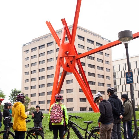 A group of people with bikes wear helmets and stand looking at Marc di Suvero's "Clock Knot," a large red sculpture that juts out over the green hill it's placed on 