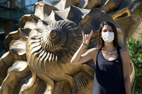 A woman in a mask makes a "hook em" gesture and looks at the camera in front of Marc Quinn's sculpture "Spiral of the Galaxy" which is an enlarged bronze seashell