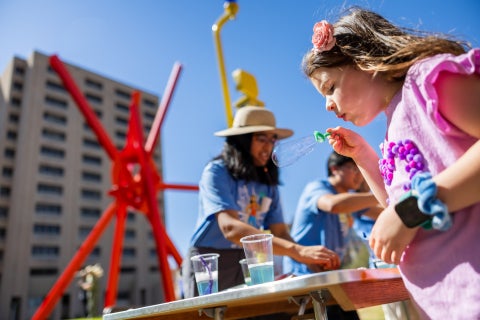 A young girl blows bubbles while in front of a large red sculpture.