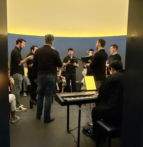 Arranged in a tight circle, a group of choral singers preform with a keyboard player.