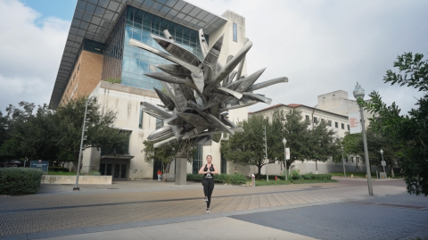 a young woman stands on one leg with her palms in a prayer position. behind her is a large sculpture made of silver canoes that is suspended in front of a building.