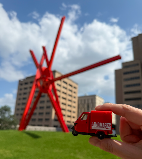 A miniature toy car with "LANDMARKS" branded on the side is held in front of a red metal sculpture