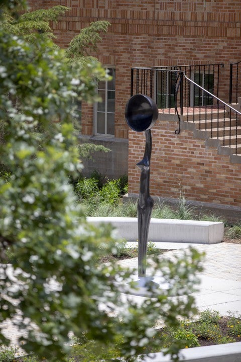 photo by Christina Murrey The artwork, Sentinel IV, a tall slender female figure with a bowlcrown for a head is seen from behind a tree branch which frames the work.