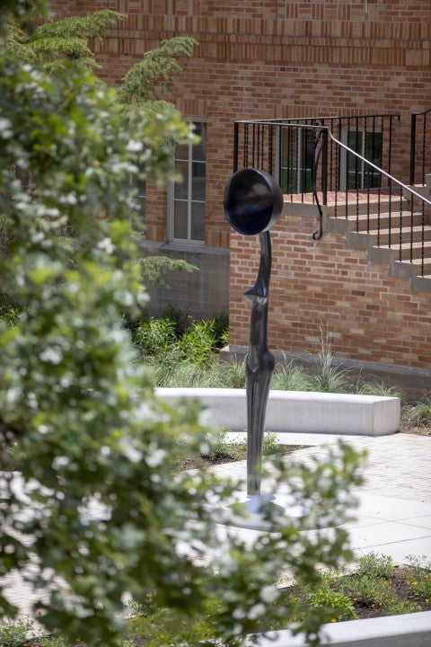 The artwork, Sentinel IV, a tall slender female figure with a bowlcrown for a head is seen from behind a tree branch which frames the work.