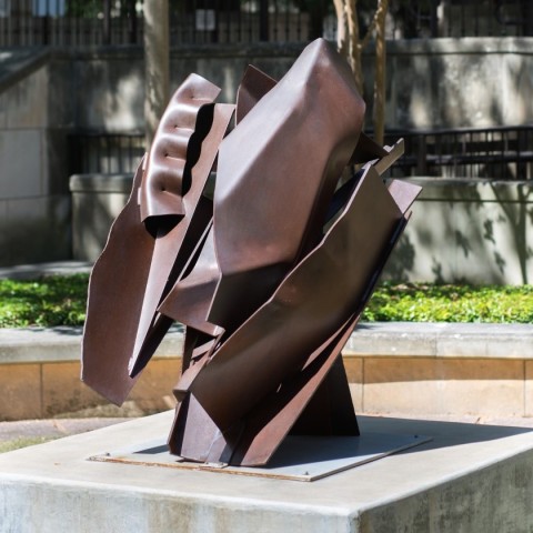 A steel sculpture that is composed of many welded together parts, the work is a dark brown. 