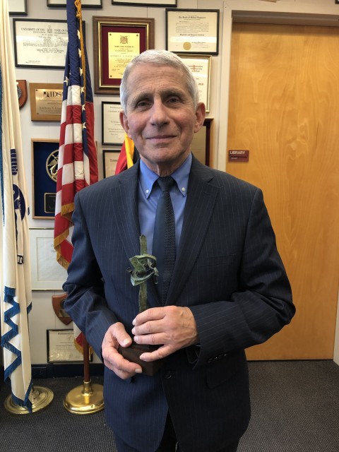 Photo shows older gentleman (Dr. Fauci) holding a small replica of Seymour Lipton's "Pioneer" in his hands. He stands in an office with an American flag in the background